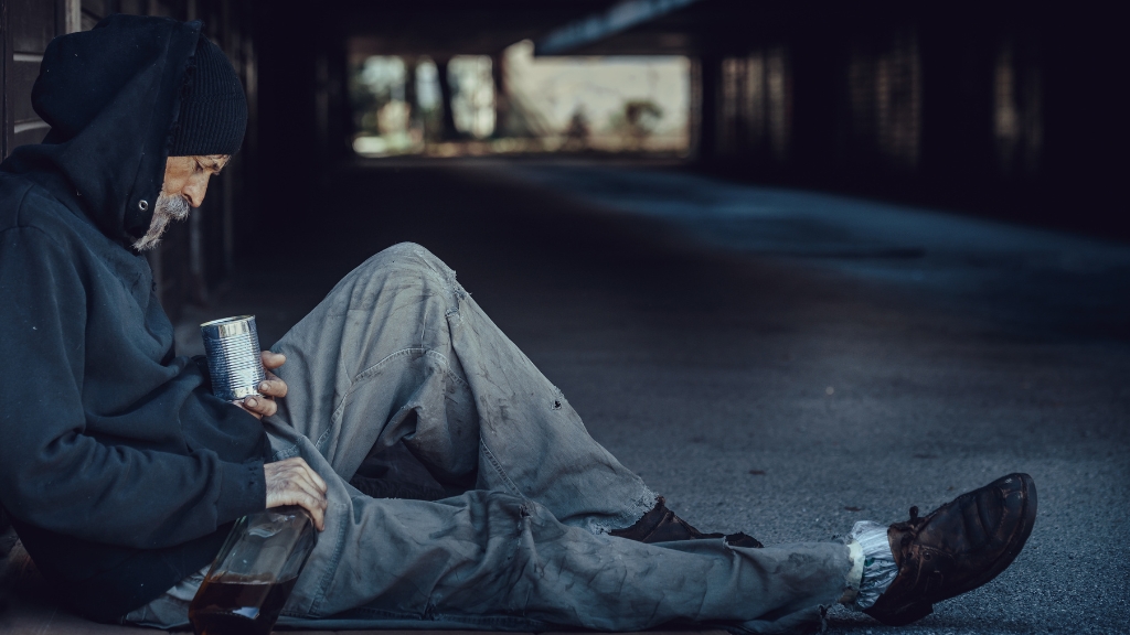 An individual sits on the ground in a dimly lit space, holding a can and bottle, representing the reality of high homeless populations