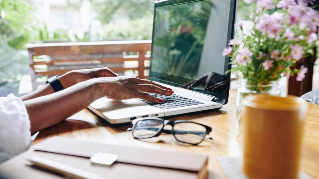 Person typing on a laptop at an outdoor desk surrounded by flowers and notebooks, illustrating remote work in a relaxed setting
