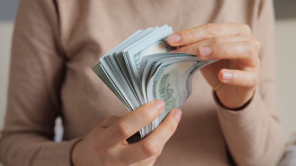 A woman wearing a beige sweater counting a large stack of hundred-dollar bills, showing income differences
