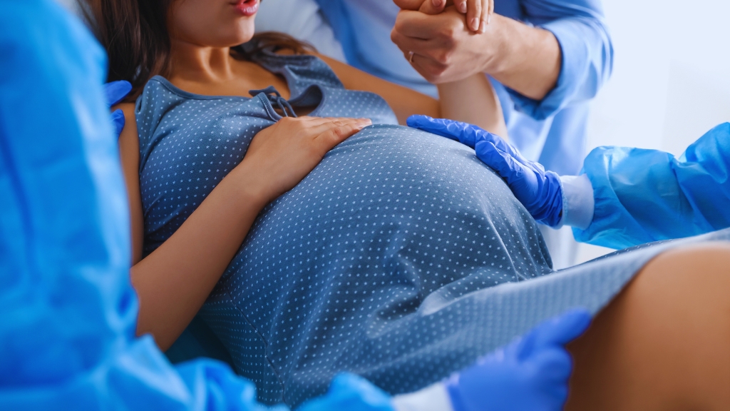 A woman in labor holds a partner’s hand while medical staff assist her childbirth in a hospital delivery room