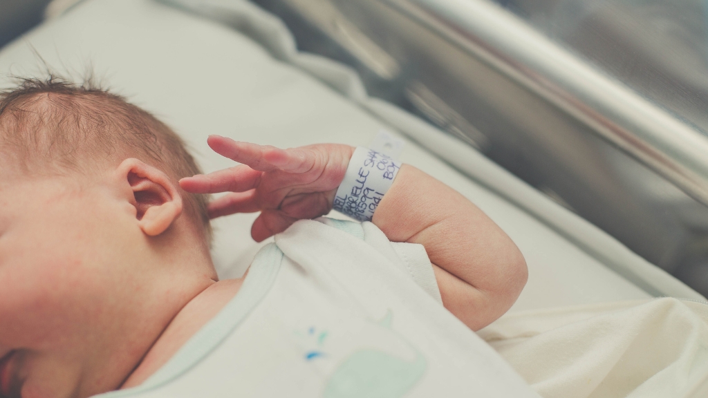 A newborn baby with a hospital ID bracelet rests in a crib, showing the reality of being born in the US