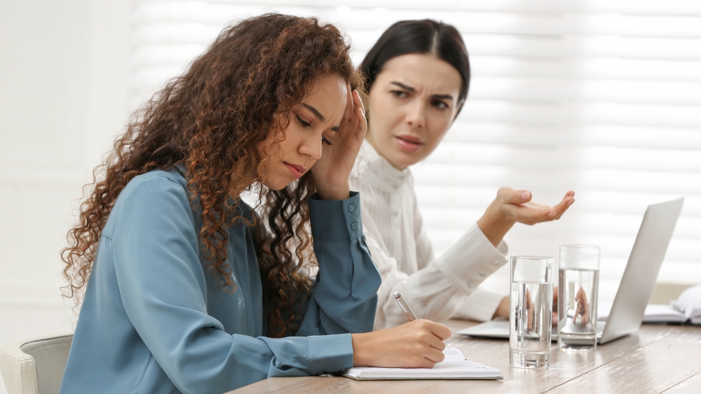 Two women sit at a desk with laptops, one looking frustrated and the other appearing confused during a discussion