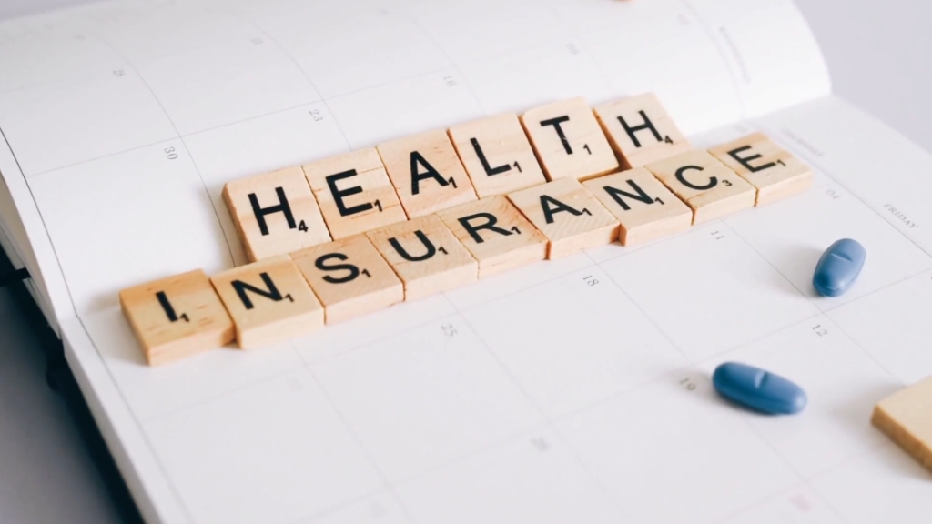 Wooden Scrabble tiles spelling “Health Insurance” placed on a calendar with two blue pills nearby
