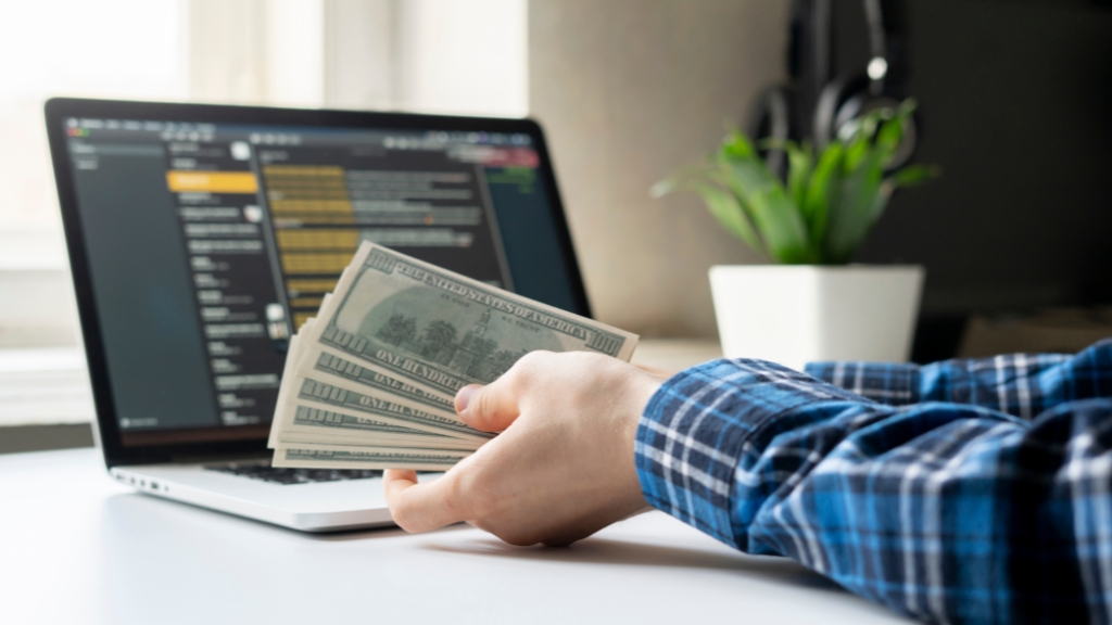 A person holds US dollar bills in front of a laptop, representing income earned through digital work
