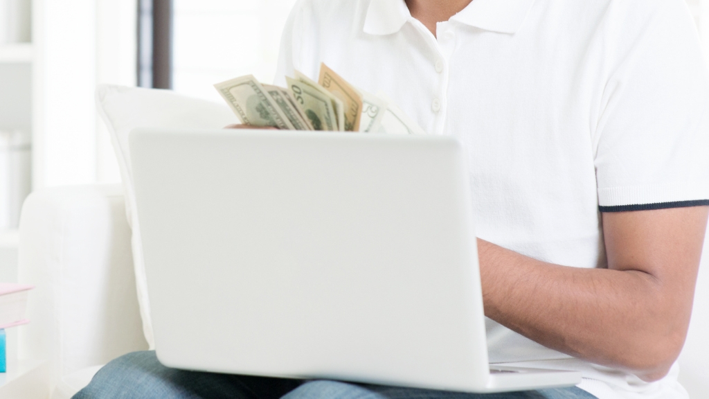 A person sits with a laptop and holds cash, symbolizing the obstacles faced in earning money online