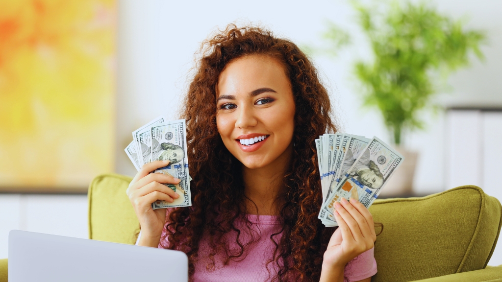 A young woman smiles while holding stacks of US dollar bills in both hands, sitting indoors with a laptop in front of her