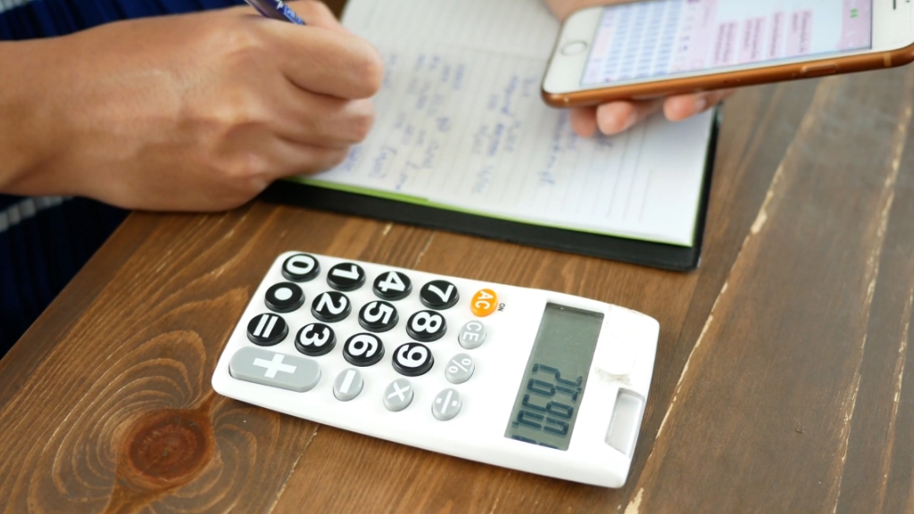 A person calculates household expenses with a calculator, notebook, and phone