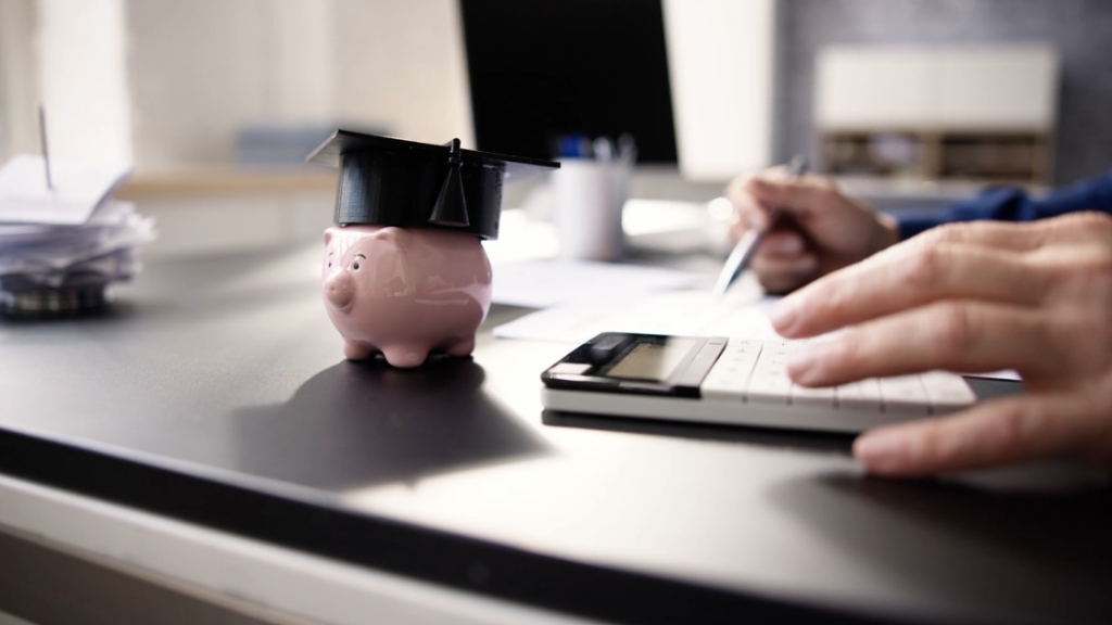 Piggy bank with graduation cap beside a person using a calculator for student loan costs