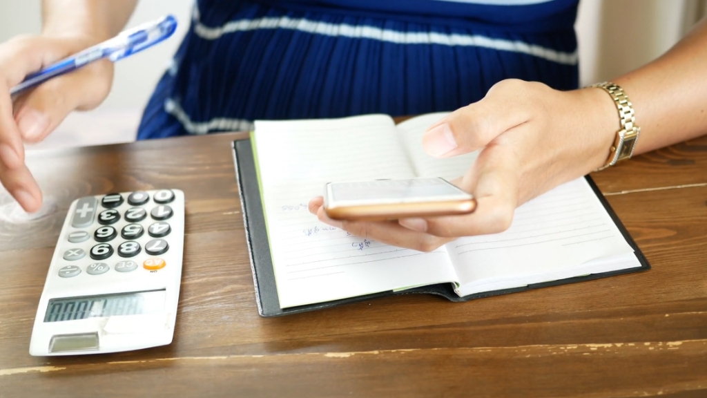 A person uses a phone, notebook, and calculator to manage utilities in the context of household budgets