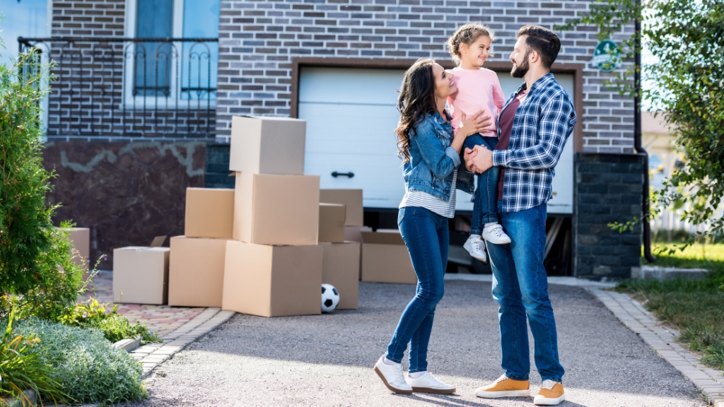 A young family stands outside their new home surrounded by moving boxes, preparing for relocating to the US for work