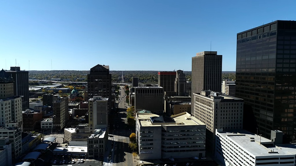 Aerial view of downtown Dayton, Ohio, with tall office buildings, mid-rise structures, and surrounding neighborhoods under a clear blue sky