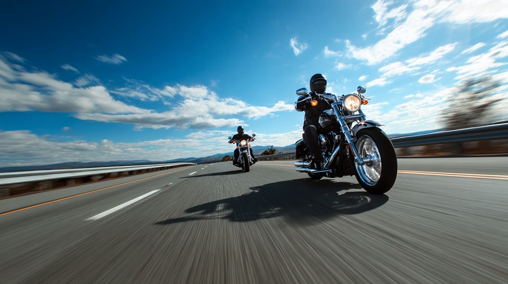 Two motorcyclists in full gear riding on a highway under a bright blue sky, with the lead rider in sharp focus
