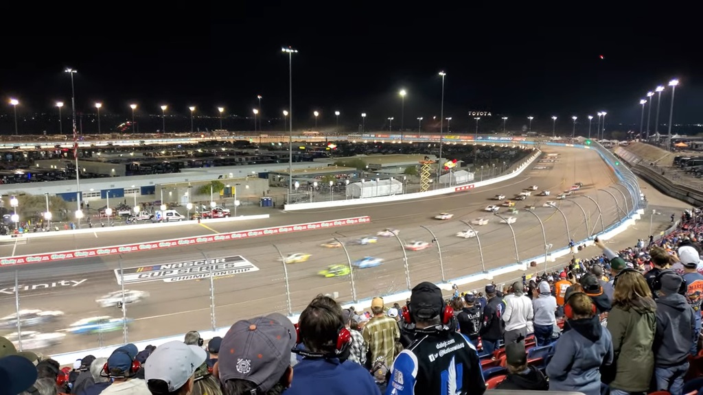 Crowd watches a night NASCAR race at Phoenix Raceway, with cars speeding around the track under bright lights