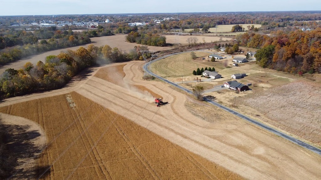 Aerial view of Kentucky farmland during harvest season, showing a combine harvester working across golden fields near rural homes and tree-lined roads