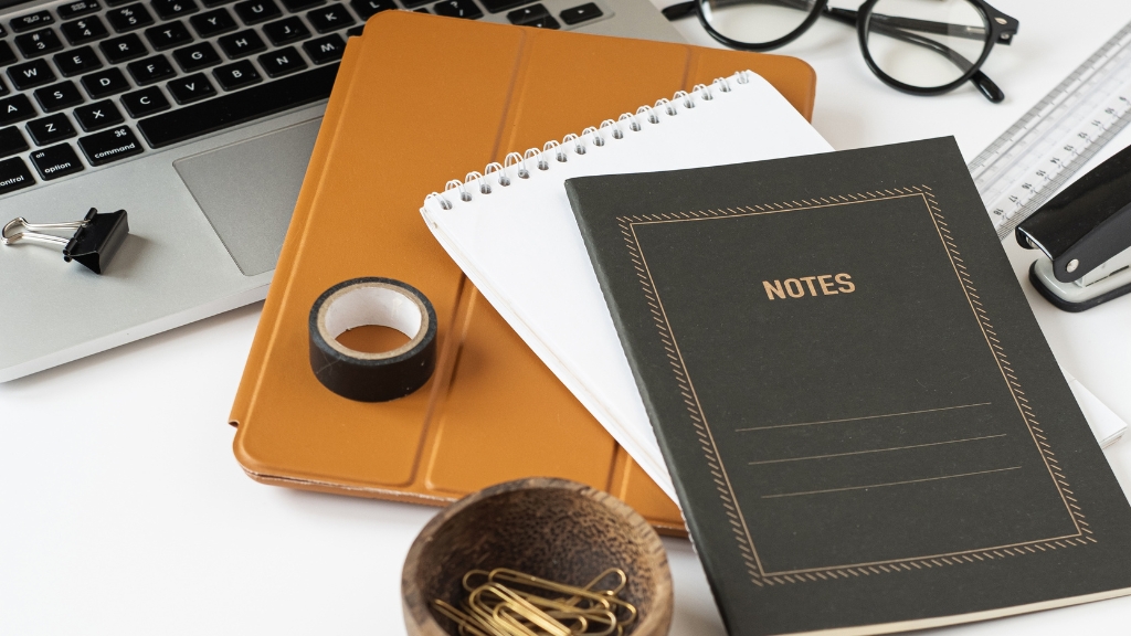A desk setup with notebooks, laptop, tape, paper clips, and office supplies arranged neatly