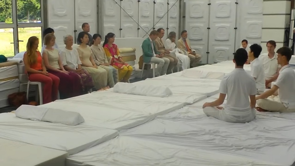 A group of people sit on white mats inside a bright hall, meditating in silence while others observe, showing a peaceful communal meditation session