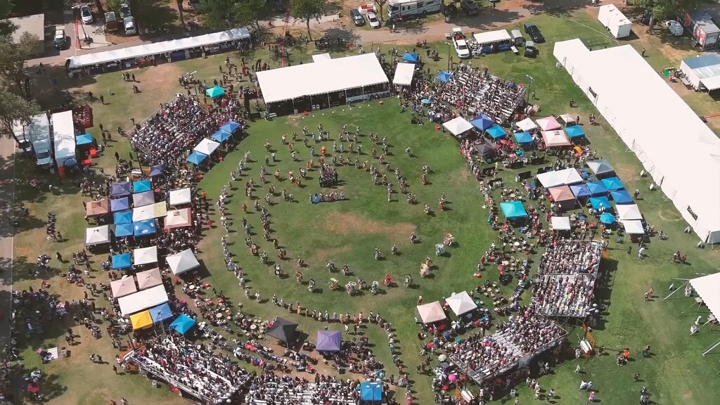 Aerial view of a large powwow gathering with dancers in concentric circles and spectators surrounding the arena