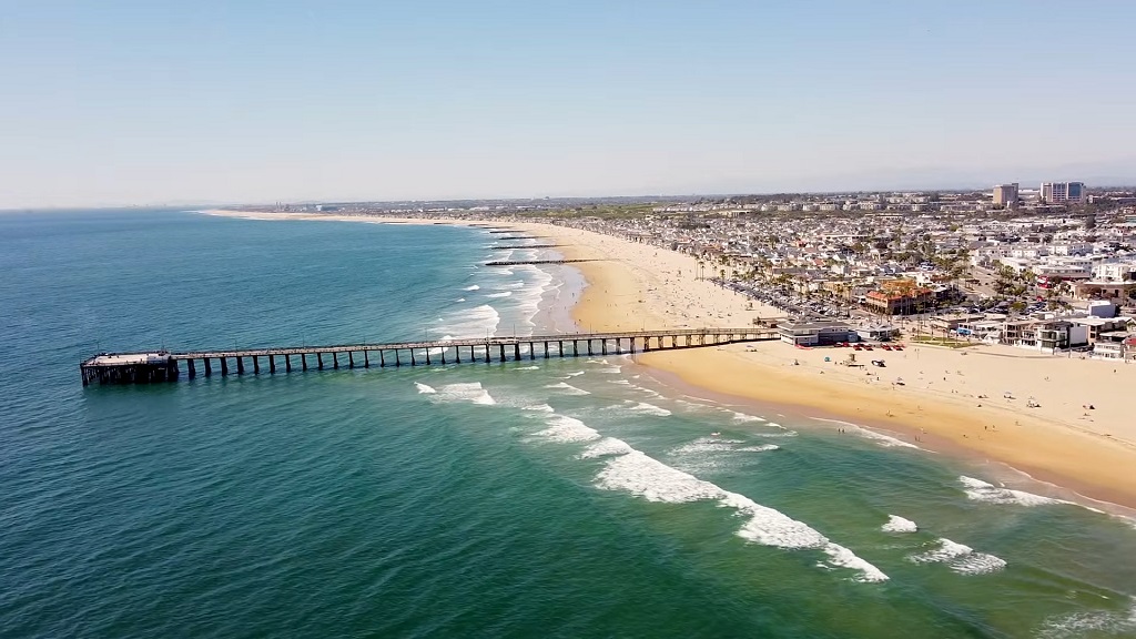 A long sandy beach with ocean waves and a pier extending into the water, bordered by a coastal city