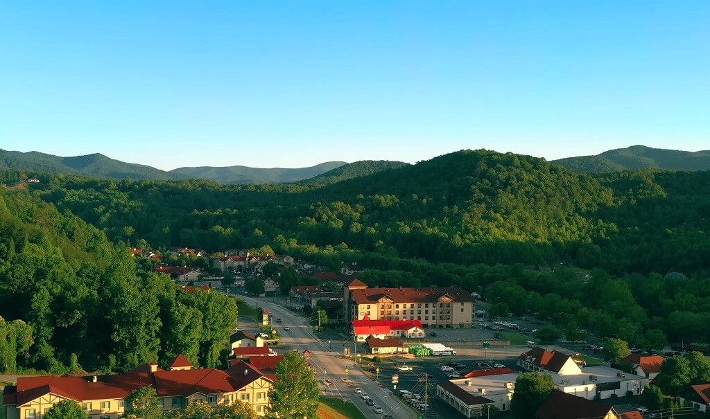 A small town surrounded by densely forested hills at sunset, with buildings and roads visible in the valley below