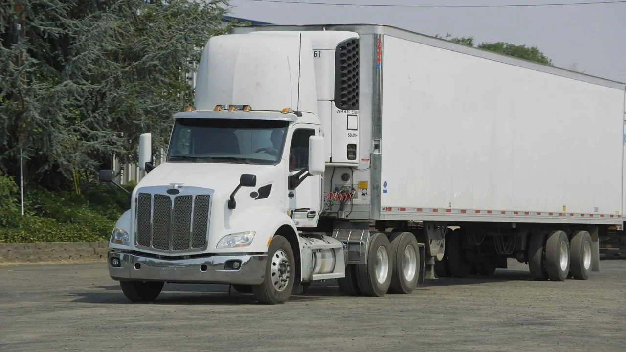 A large white freight truck driving near an industrial area, representing cross-border trade and transportation between the U.S. and Canada
