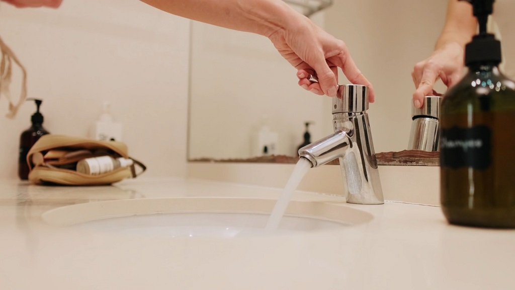 Hand turning on a bathroom sink faucet with running tap water, representing everyday household water use