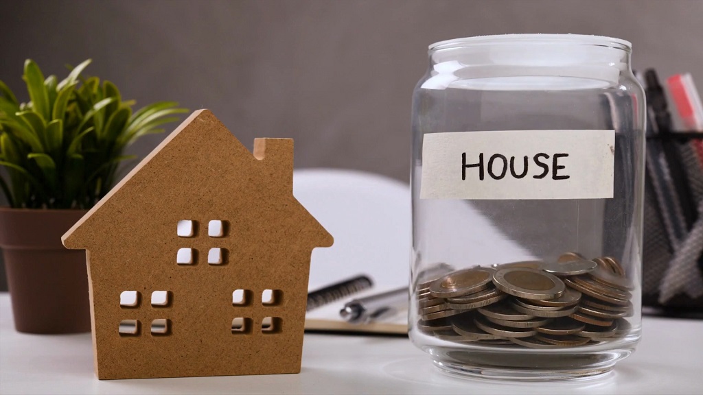 Wooden house model placed beside a glass jar labeled “House” filled with coins, symbolizing savings, housing costs, and the financial barriers to home ownership