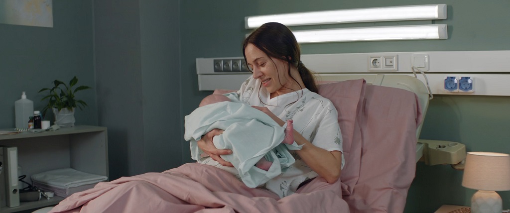 A new mother rests in a hospital bed while holding her newborn baby, smiling during post-delivery recovery