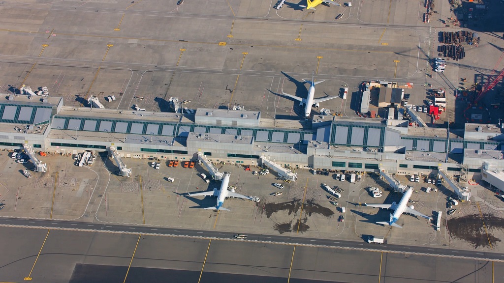 Aerial view of multiple commercial aircraft parked at terminal gates, with jet bridges, service vehicles, and airport apron visible