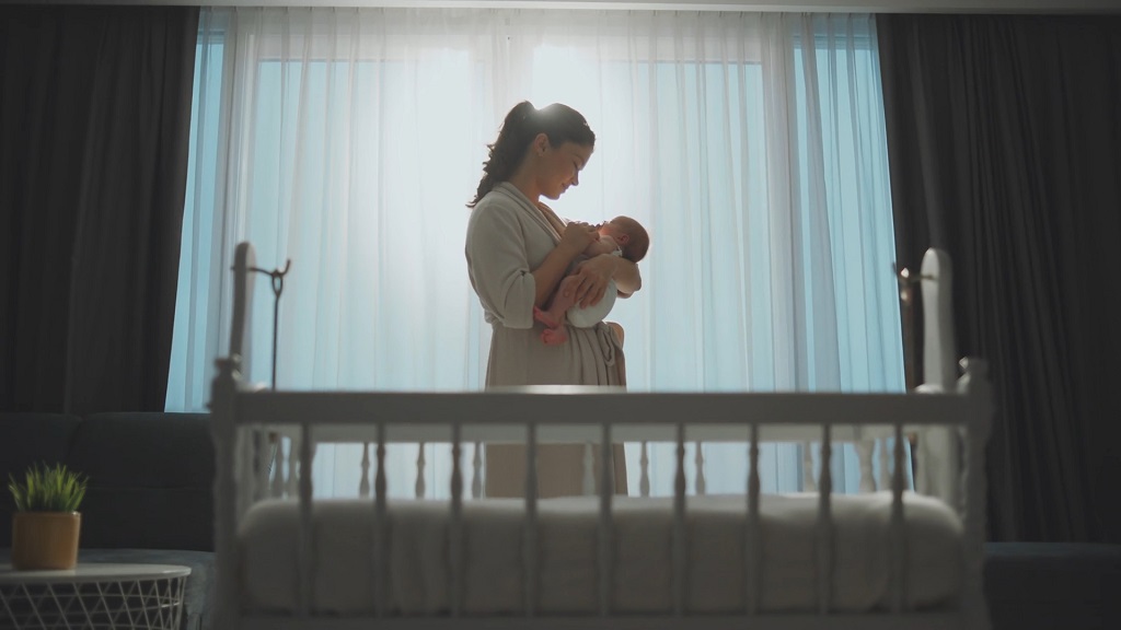 A mother stands in a softly lit room holding her newborn baby near a crib, highlighting the early bonding period after birth