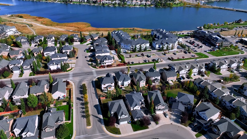 Aerial view of a modern suburban community in Alberta with neatly arranged houses, wide roads, and a large lake surrounded by residential and commercial buildings