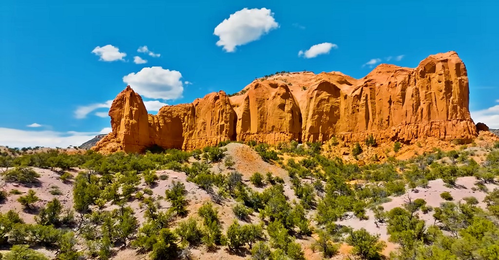 Red sandstone cliffs rising above a desert landscape with sparse green shrubs under a bright blue sky with scattered clouds