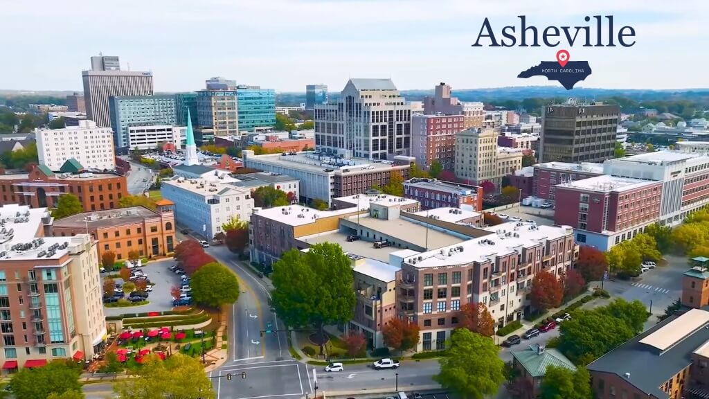 Aerial view of downtown Asheville with mid-rise buildings, tree-lined streets, and surrounding mountain foothills