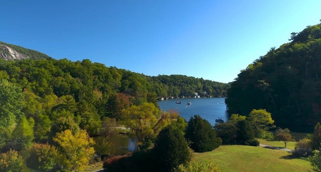 A calm lake bordered by green forested hills on a clear sunny day, with boats visible on the water