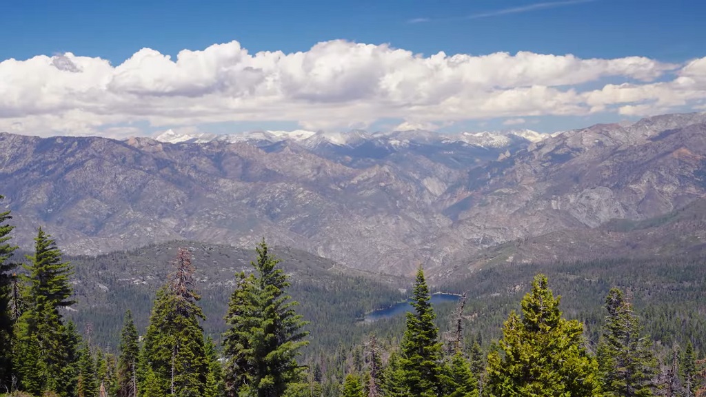 Wide panoramic view from Panoramic Point in Kings Canyon National Park, showing layered Sierra Nevada mountains, forested slopes, distant snowcapped peaks, and a clear blue sky
