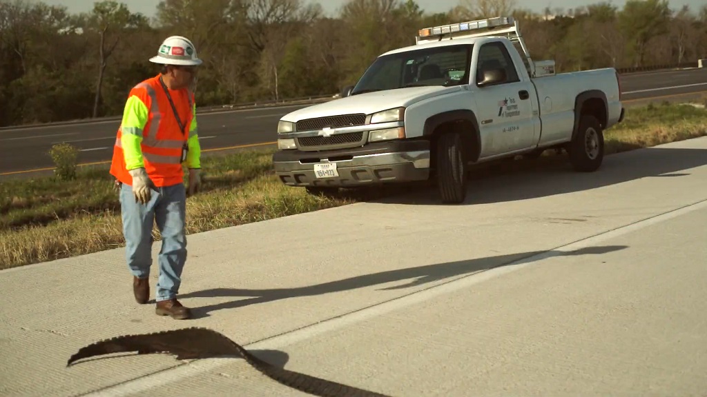 Roadside worker in high visibility safety vest inspecting pavement damage beside a parked utility pickup truck on a Texas highway shoulder