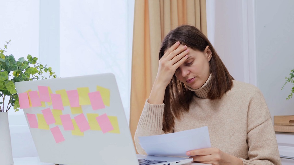 Woman sitting at a desk reviewing bills and paperwork beside a laptop covered with sticky notes, conveying financial stress and budgeting concerns for renters
