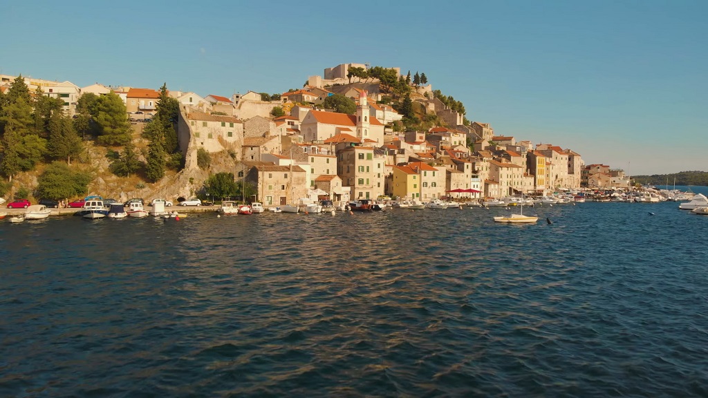 Coastal view of Šibenik, Croatia, with historic stone buildings on a hillside and small boats docked along the waterfront
