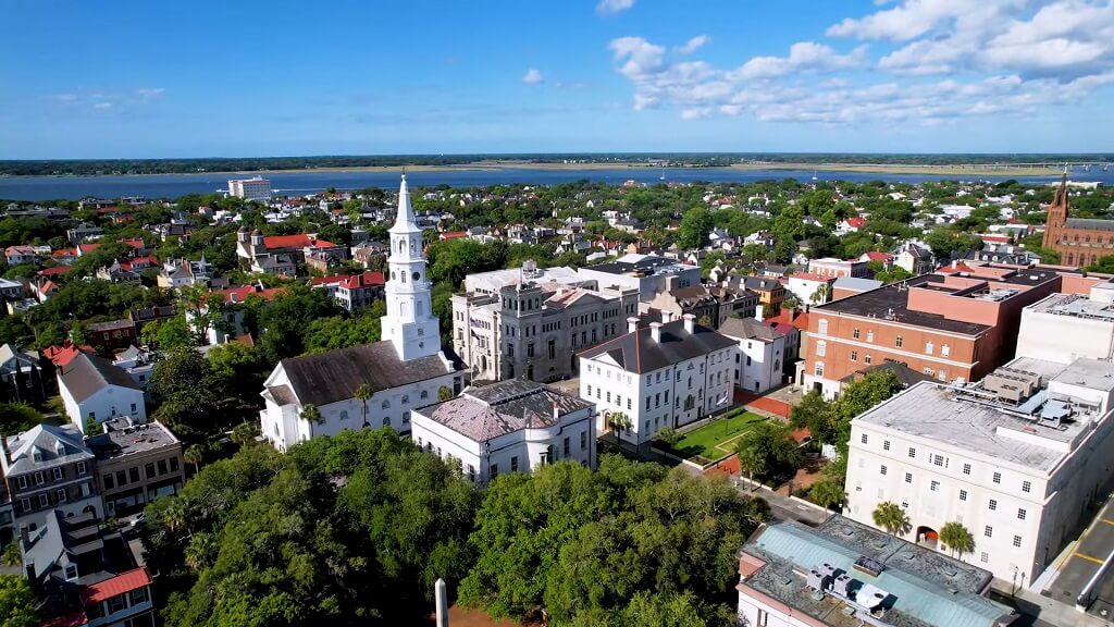 Aerial view of a historic city center with church steeples, leafy streets, and waterfront in the distance under a clear sky