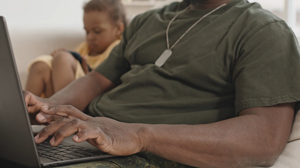 Veteran working on a laptop at home while a child sits nearby, representing education and career training after military service