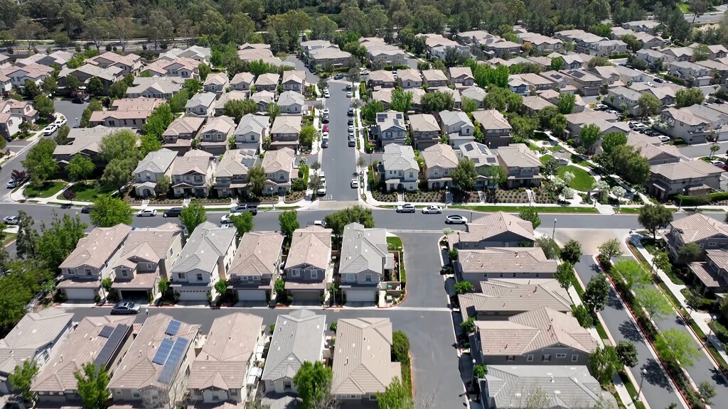 Aerial view of a suburban residential neighborhood in California, showing rows of closely spaced single-family homes with tree-lined streets and parked cars, illustrating dense housing development